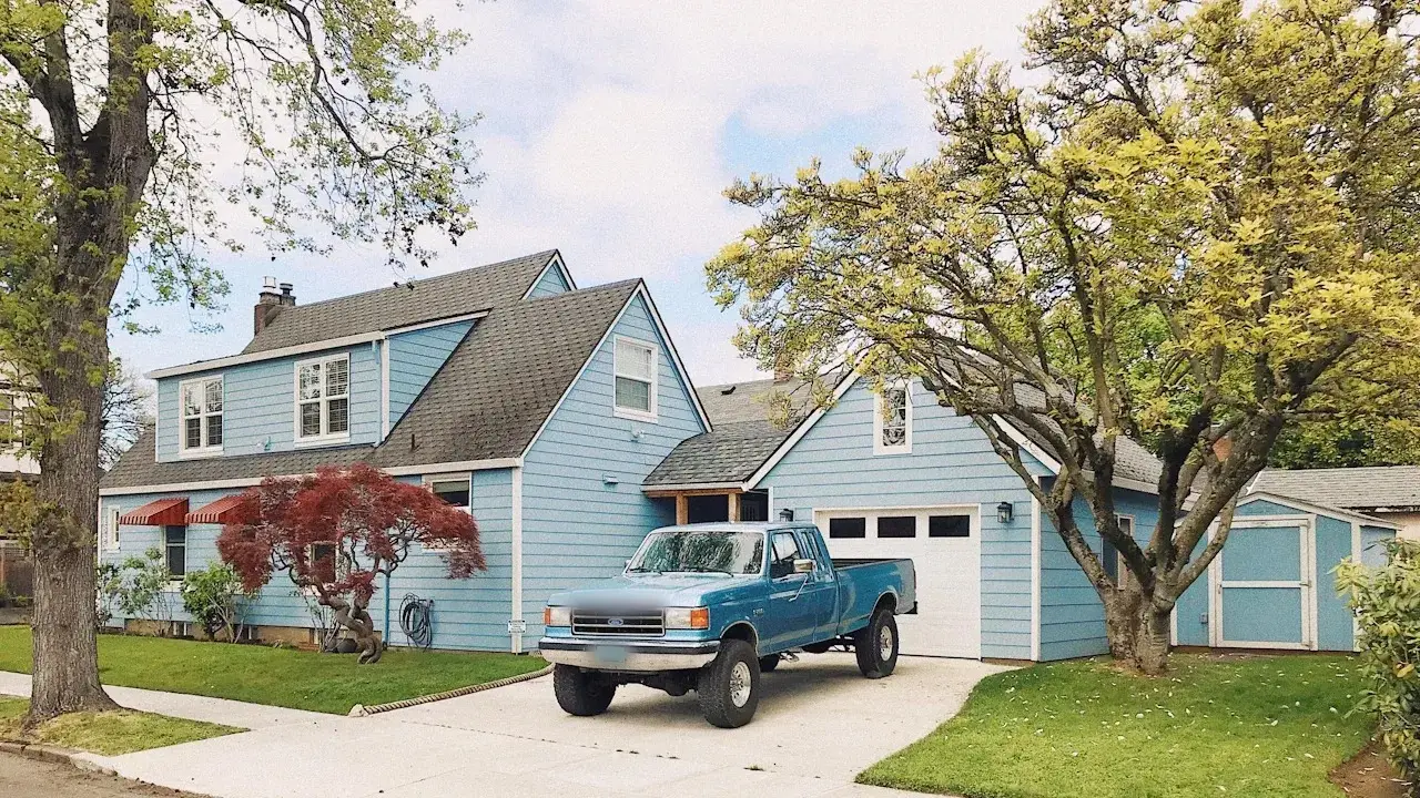An older blue pickup truck parked on the driveway of a pale blue house with a garage, under spring trees.