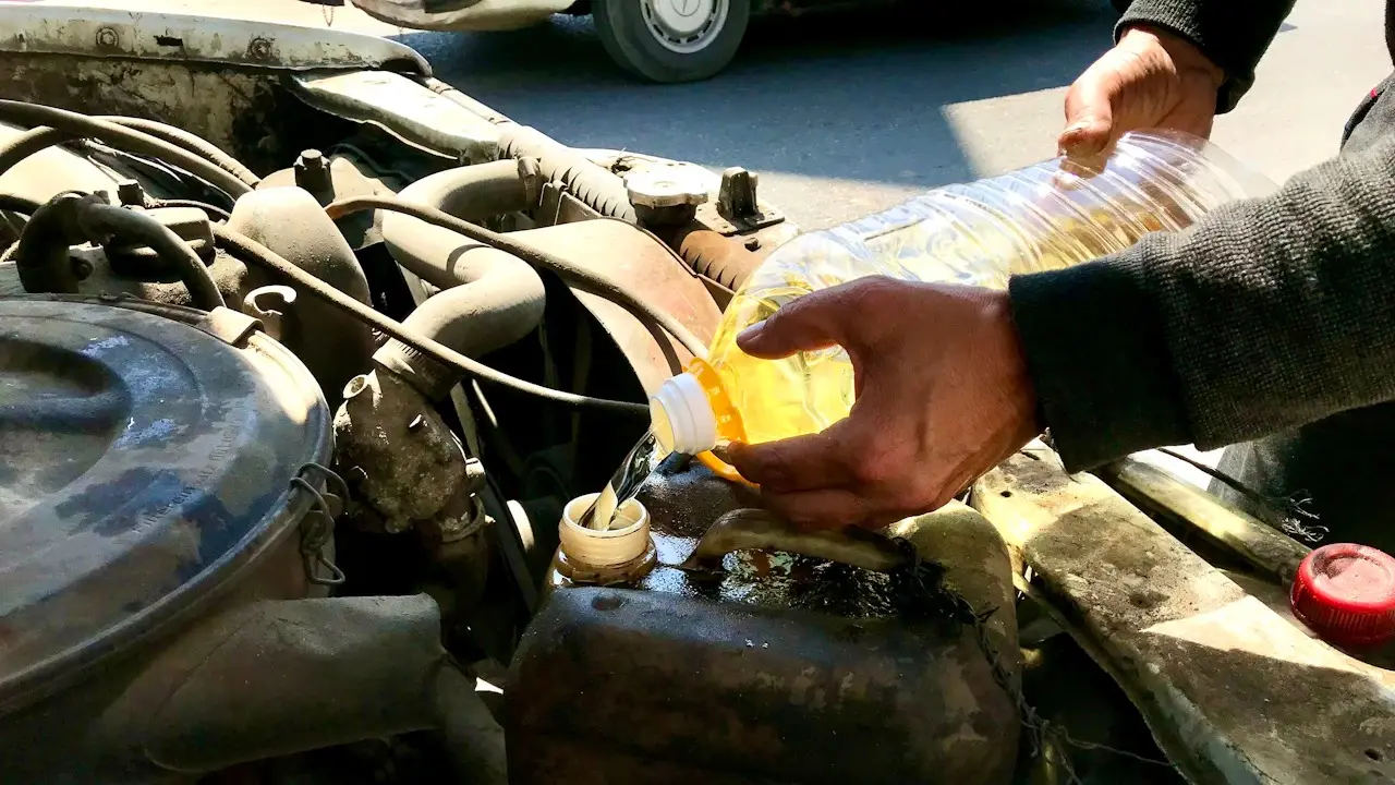 A hand pouring oil from a clear bottle into an older engine's filler neck.