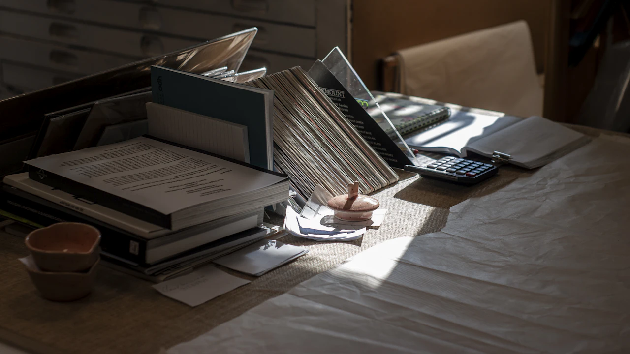A dim desk in a shaft of light: a leaning stack of thick books and bound reports, an open laptop, a desktop calculator in a document tray, loose papers and a small cup, all on a pale cloth-covered table.