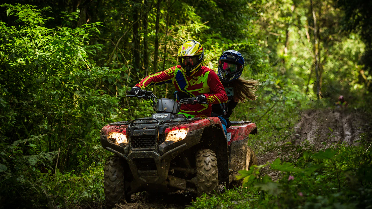 Two helmeted riders on a red quad bike coming through mud and standing water on a narrow forest trail
