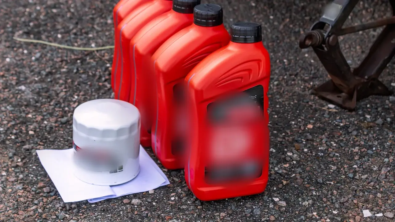Four quart bottles of motor oil and a new oil filter on gravel beside a jack.