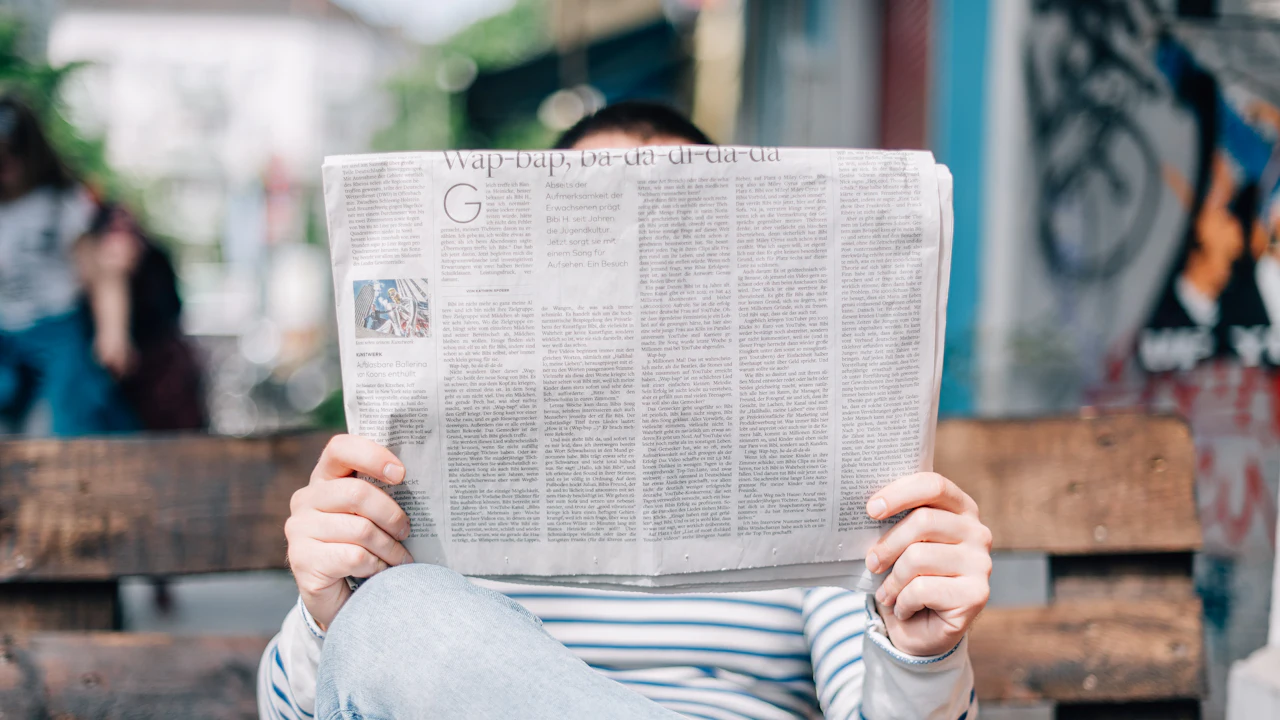 A person sitting on a bench, seen from the front, holding an open broadsheet newspaper up so it hides their face, blurred greenery and a wall behind them.