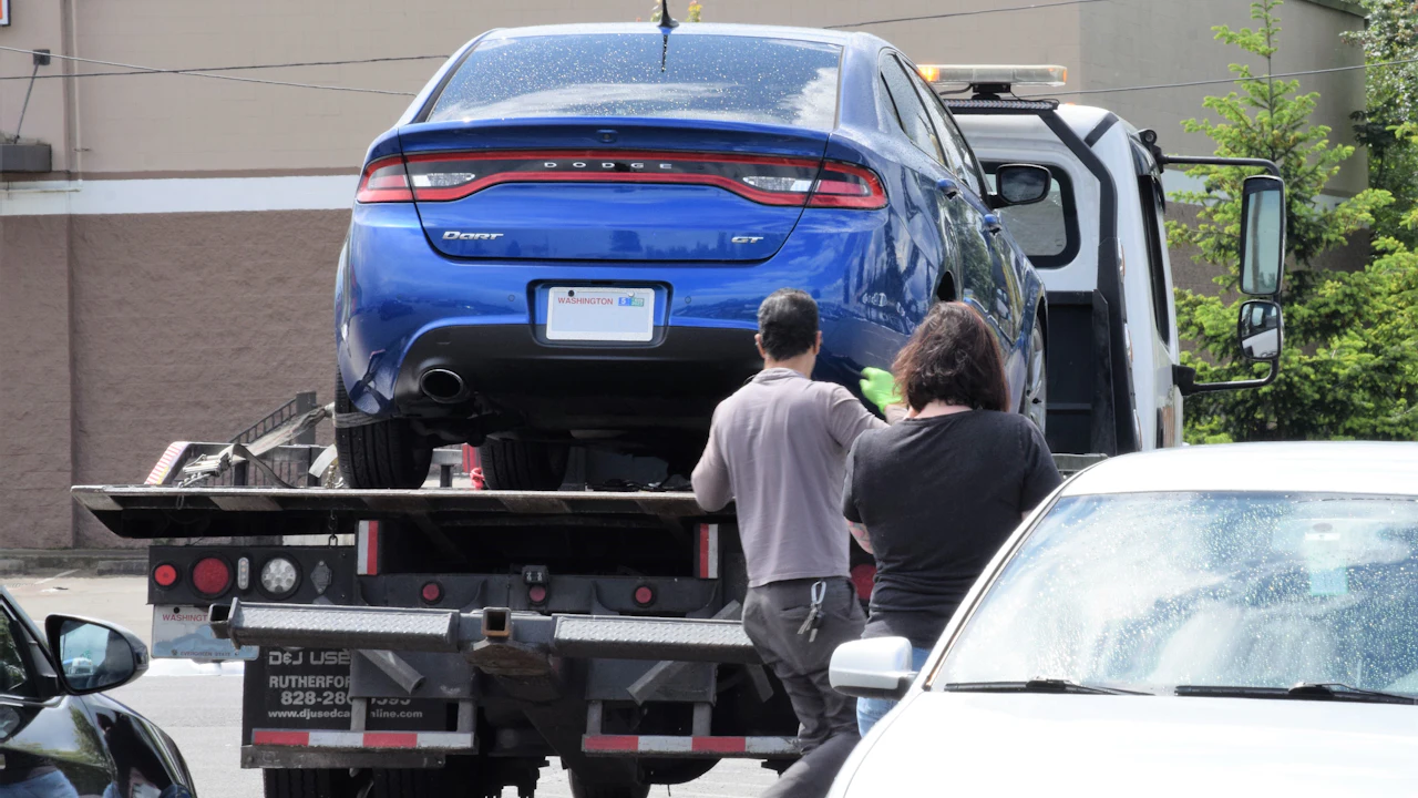 A blue saloon loaded onto a flatbed tow truck while two people stand watching beside it