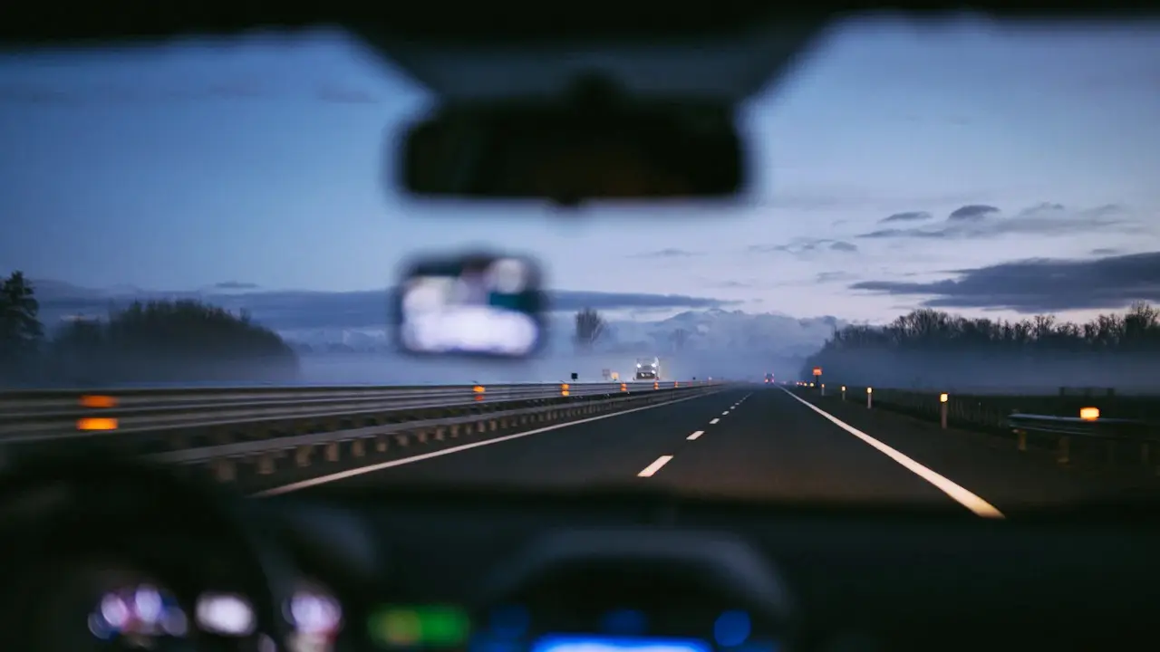 A motorway at dusk seen from the driver's seat, the dashboard lights and rear-view mirror blurred.