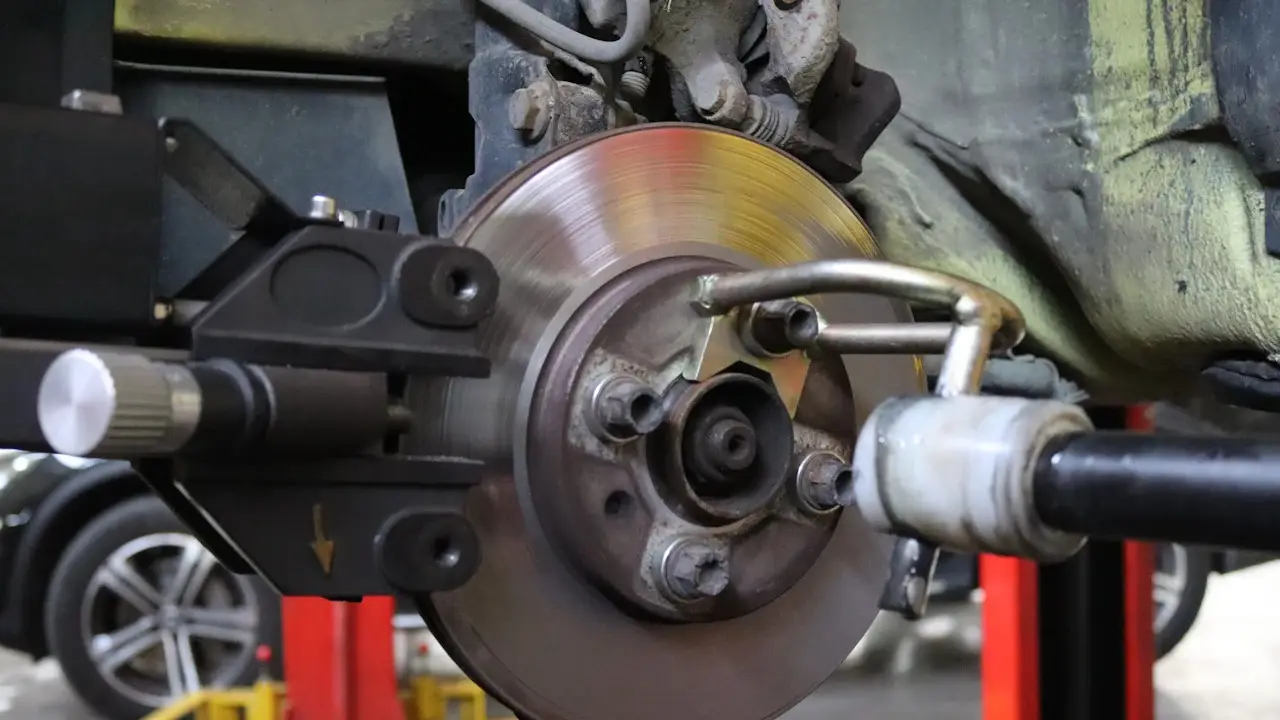 A car’s brake corner on a lift with the wheel off, showing the rotor, caliper bracket and brake line.