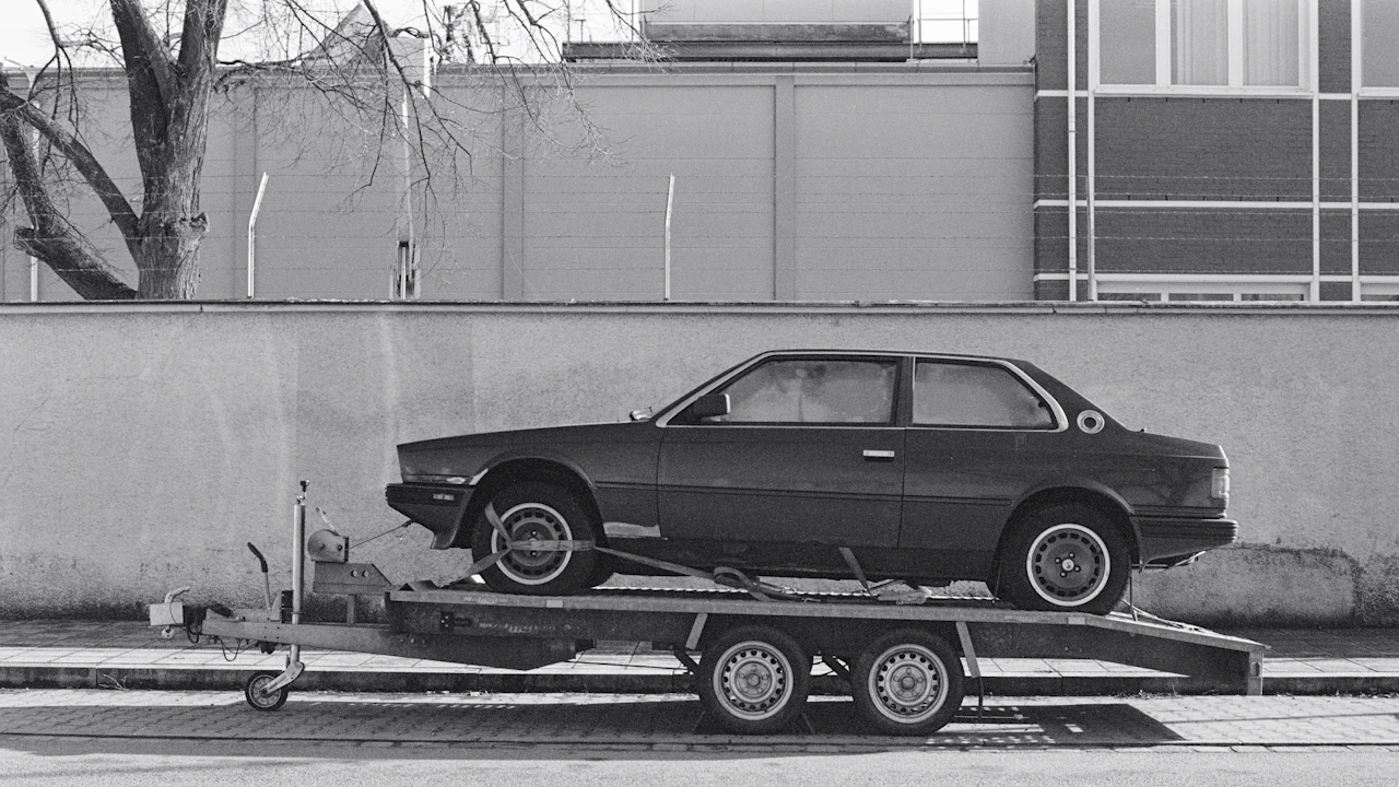 An old coupe strapped to a twin-axle trailer at a kerbside, photographed side-on in black and white