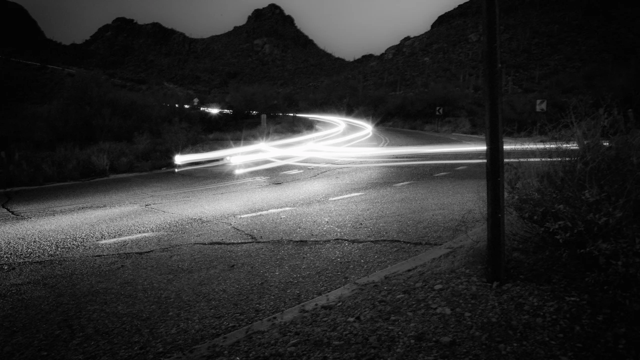 A long-exposure night shot of a desert highway, a car's light trail curving past dark saguaro hills