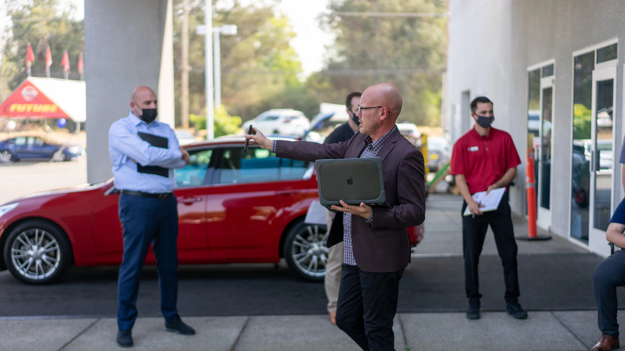 A man holding an open laptop gestures towards a red saloon on a dealership forecourt while colleagues with clipboards look on
