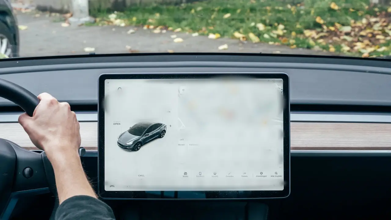 A driver's hand on the steering wheel beside a large centre touchscreen; the screen's contents blurred.