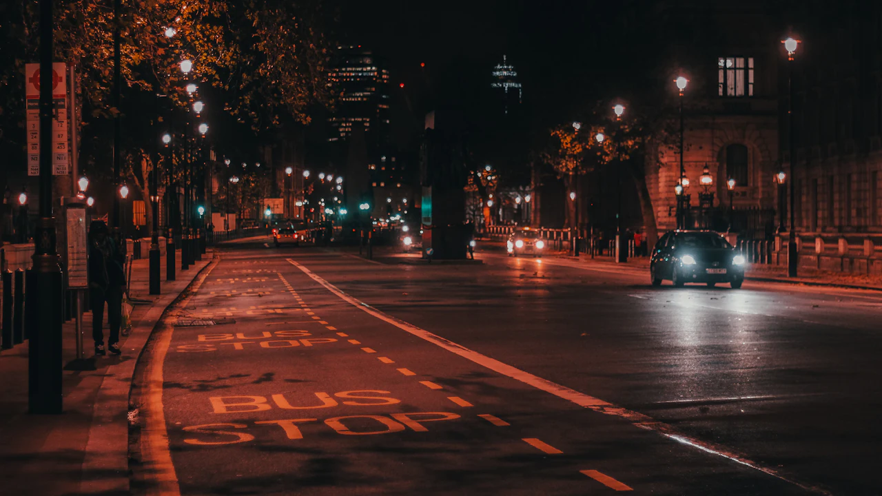 A city street at night lit by rows of orange street lamps, a dark saloon approaching with its headlights on and a bus stop marked on the tarmac