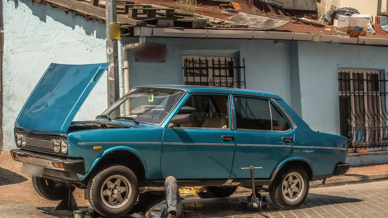 An older sedan on jack stands with its hood up and a mechanic working underneath.