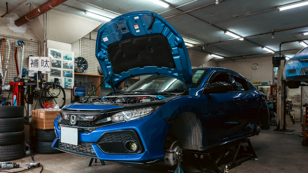 A blue hatchback with its bonnet open, raised on a workshop lift beside racks of stacked tyres