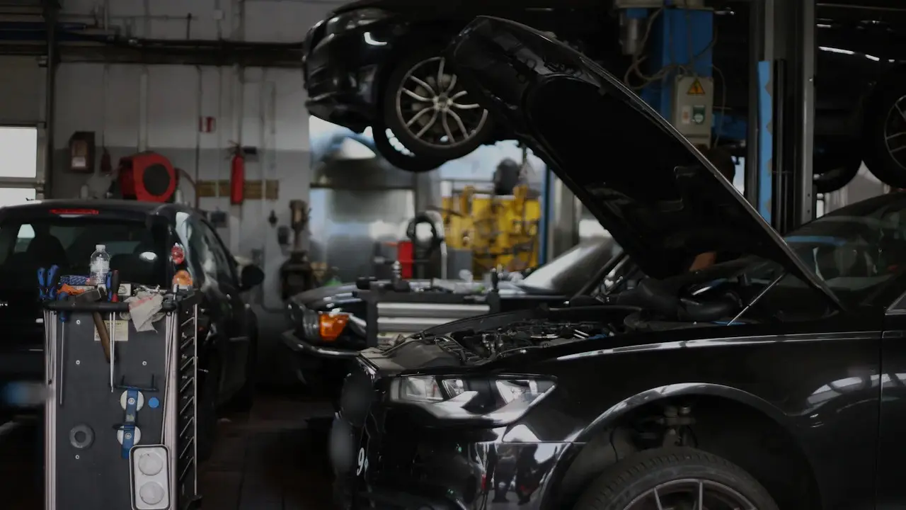 Cars in a repair shop: one raised on a lift, one with its hood open and one parked behind a tool cart.