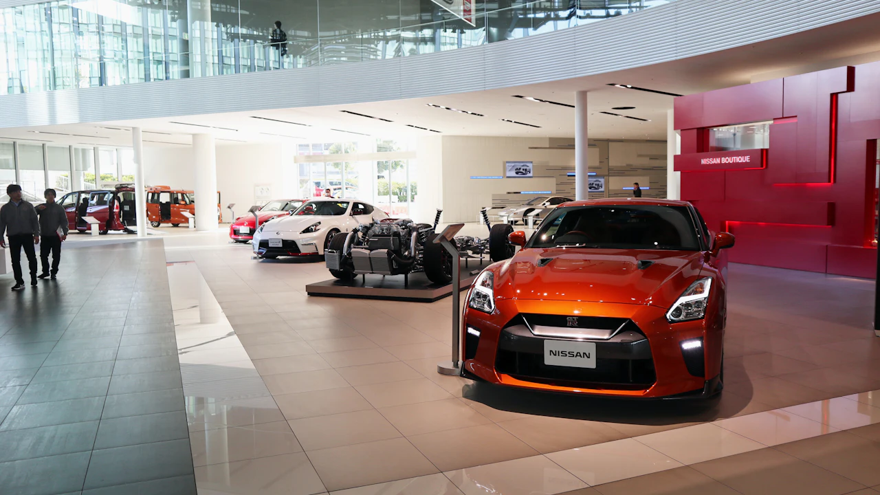 The interior of a modern franchised car showroom, an orange sports coupe displayed nose-out under a high glazed atrium with a cutaway chassis on a plinth behind it and two people walking the polished floor to the left