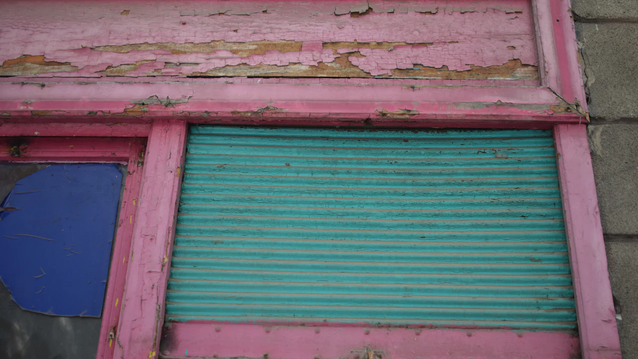 The facade of a derelict building: a peeling pink-painted frame around a corrugated turquoise roller shutter, a broken blue-painted window pane to the left, bare cinder block above.