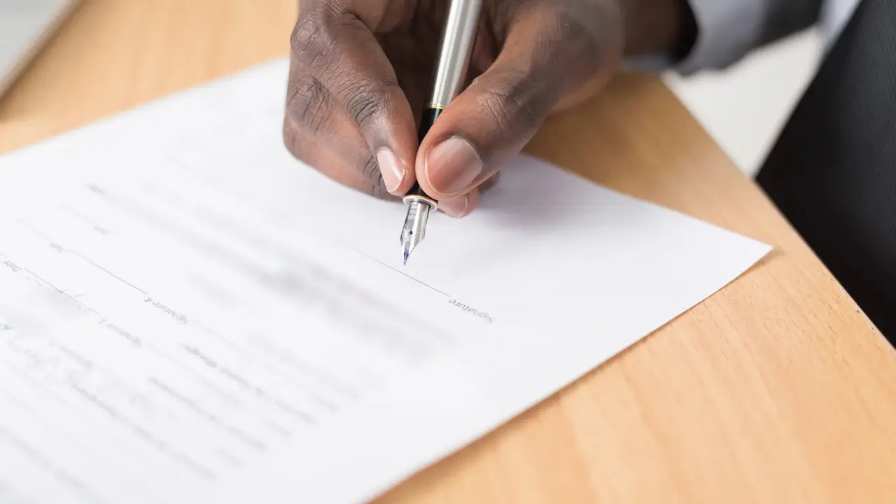 A hand signing a printed form with a fountain pen on a wooden desk; the form's text blurred.