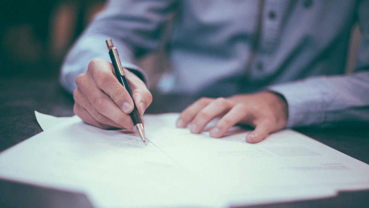 A person in a light shirt signing a printed form on a dark desk with a slim black pen