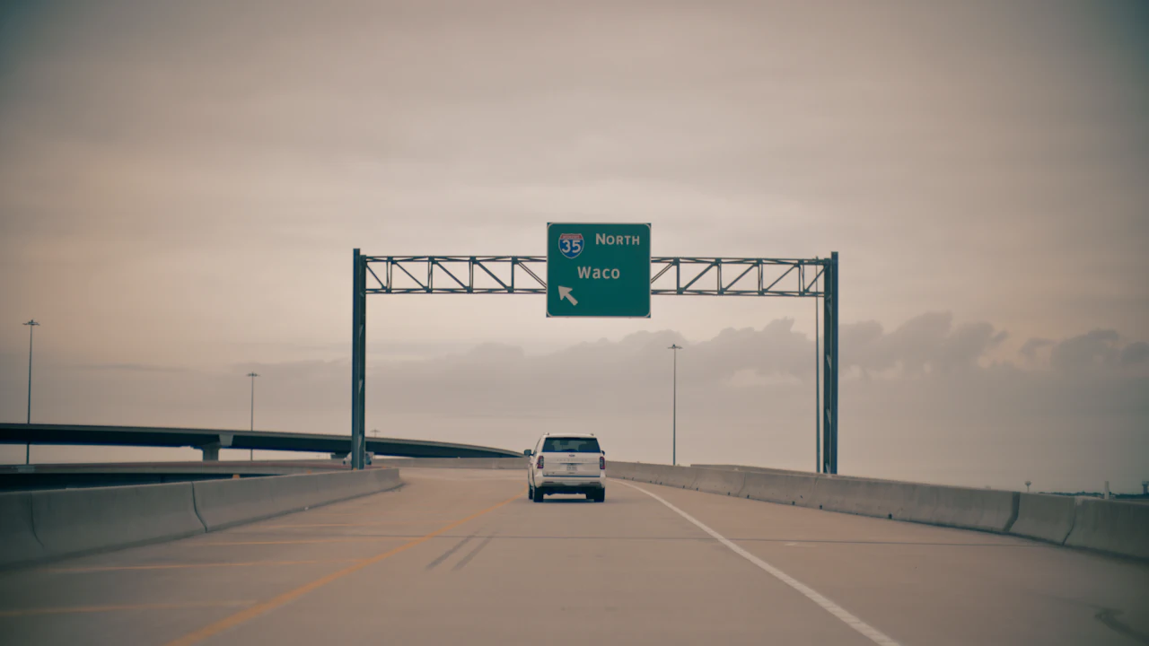 A white SUV on an empty elevated interstate under a flat grey sky, passing beneath a green overhead sign for Interstate 35 north