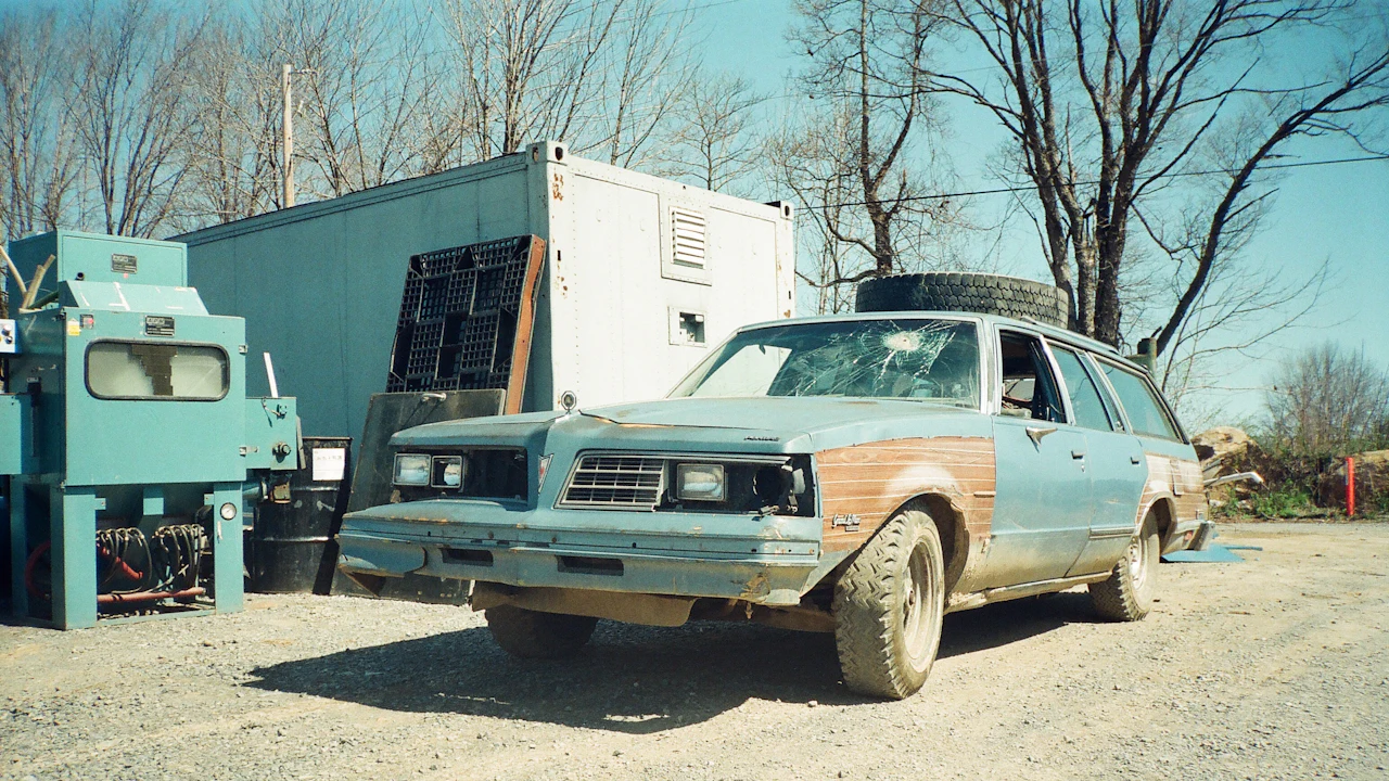 A derelict wood-panelled estate car from the nineteen-eighties standing on gravel in a yard, its windscreen shattered and one front wing a different colour from the rest