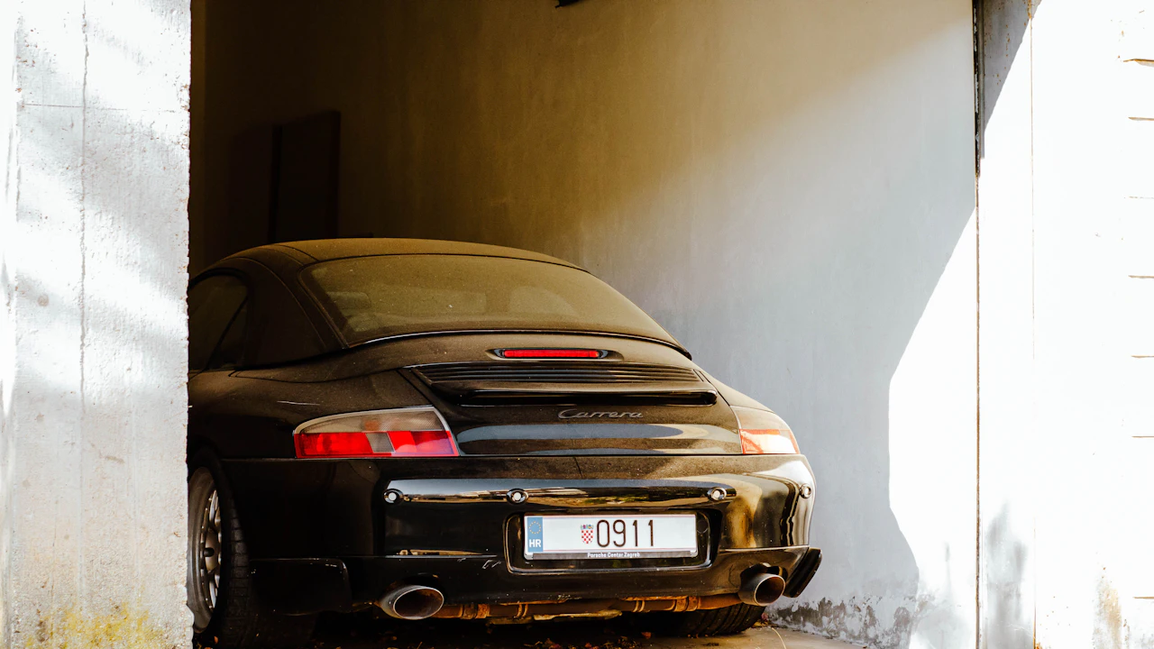The rear of a black Porsche coupe in a garage doorway, its twin exhaust tailpipes below the bumper