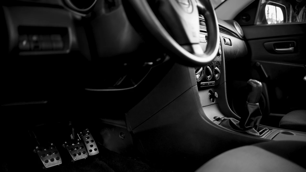 A car's footwell showing the metal accelerator, brake and clutch pedals, with the steering wheel and gear lever above
