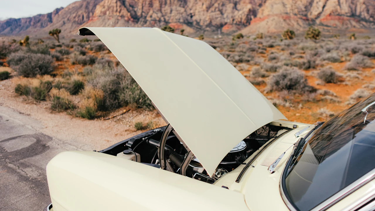 A cream car stopped on a desert roadside with its hood raised, red rock hills behind