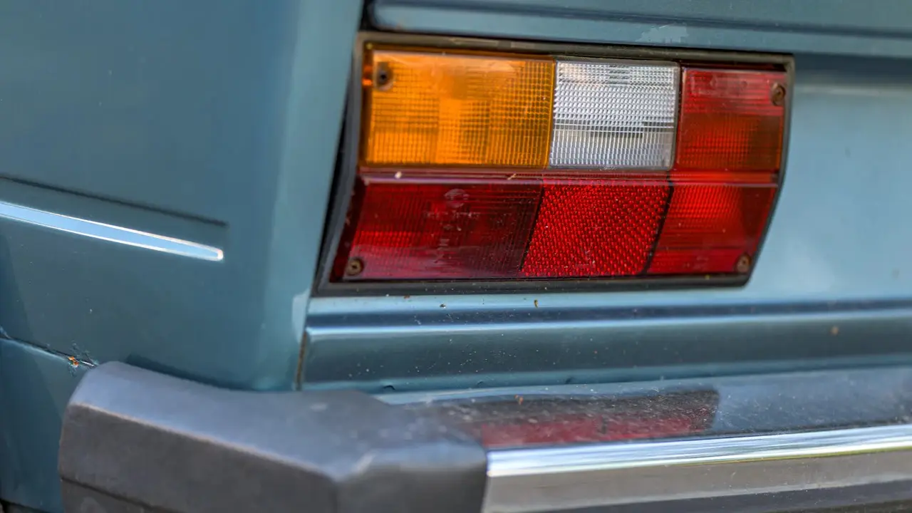 The tail light and rear bumper of an older pale blue car.