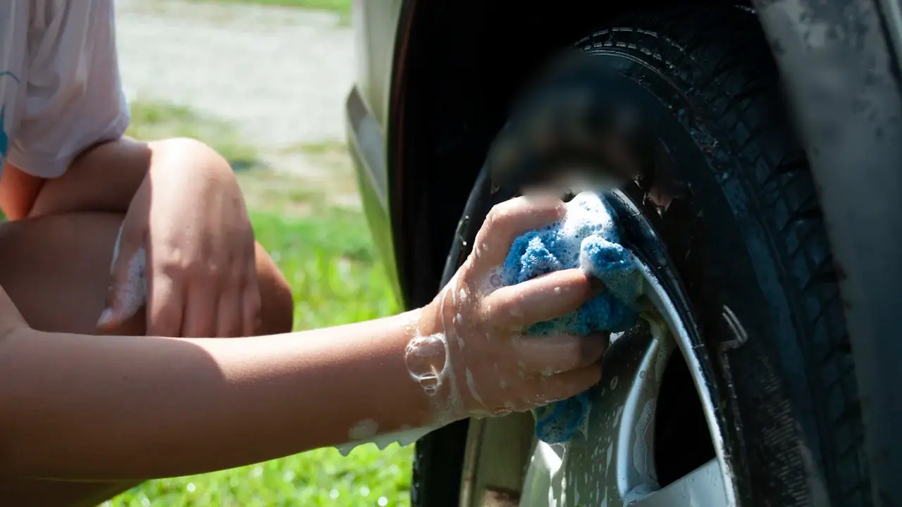 A hand scrubbing a car’s tire and wheel with a soapy sponge.