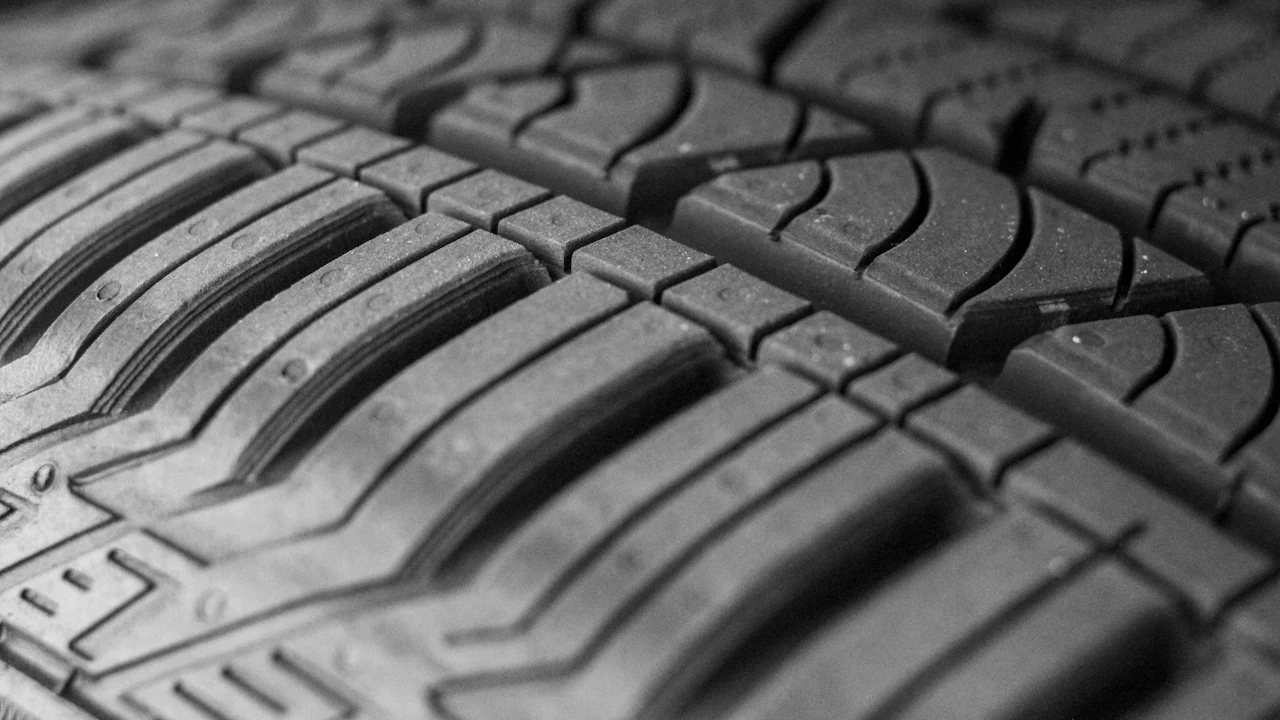 A black-and-white macro photograph of a car tyre's tread running diagonally across the frame, with deep circumferential grooves and angled blocks.