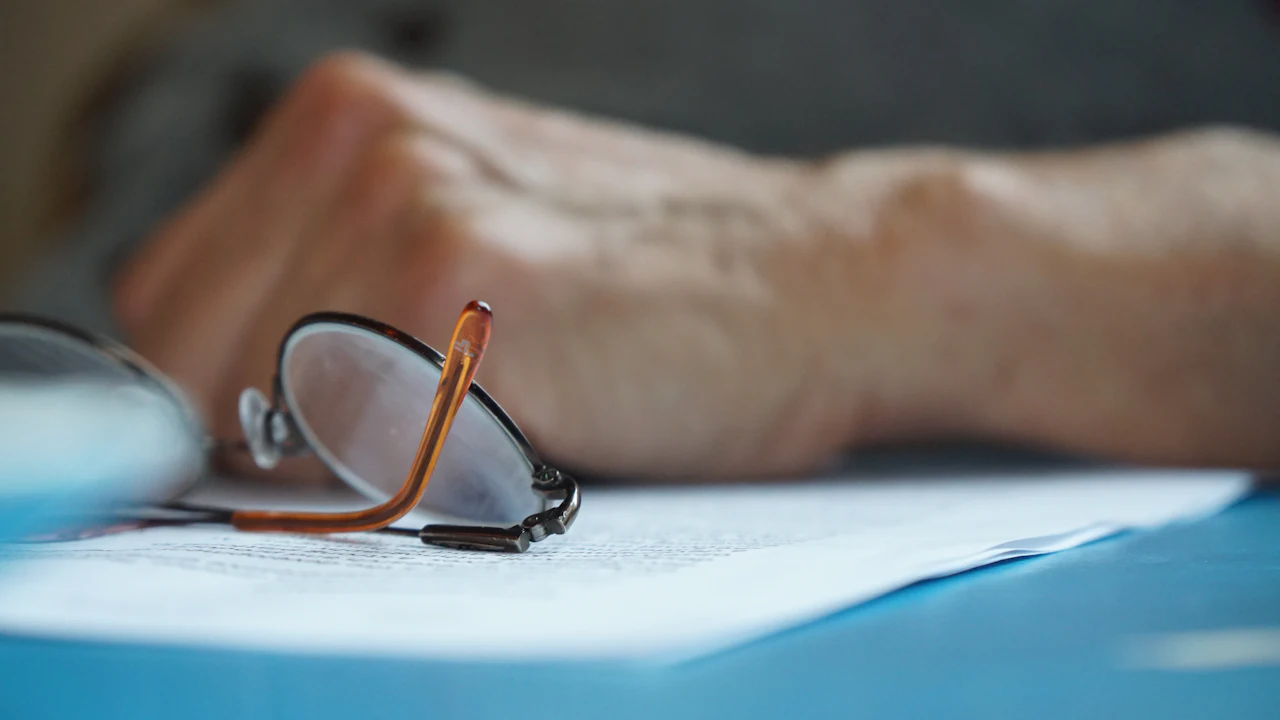 A pair of reading glasses folded on top of a printed document on a blue table, a hand resting out of focus behind them