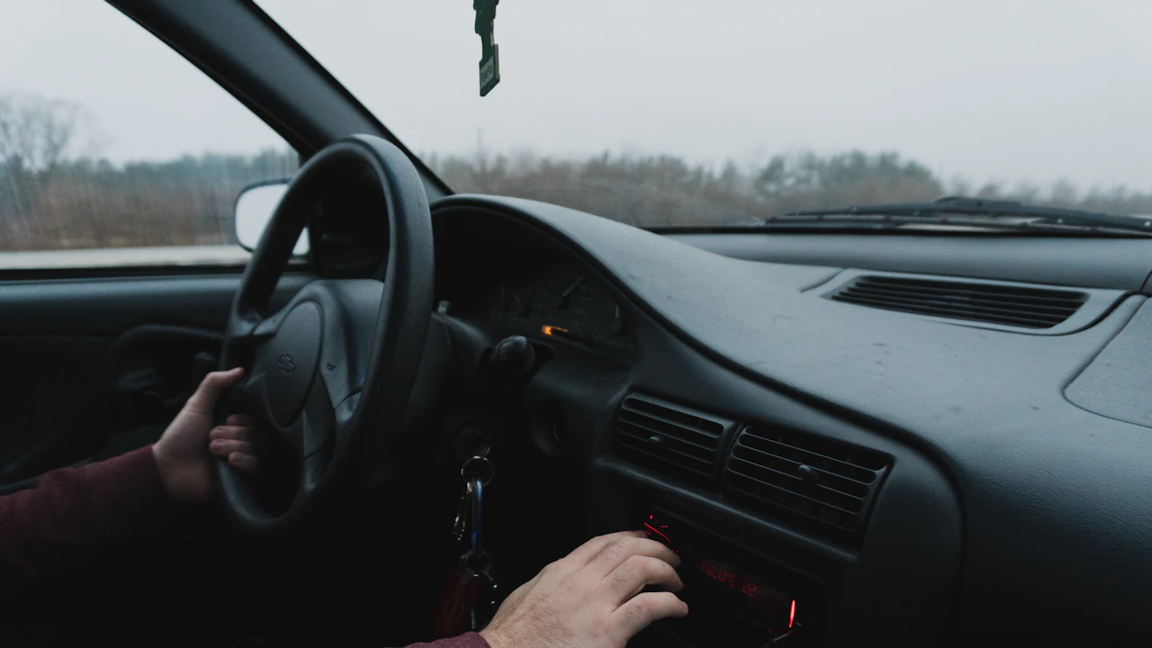 The interior of an older car seen from the passenger seat, the driver&rsquo;s hands on the wheel and the stereo, keys hanging from the ignition barrel