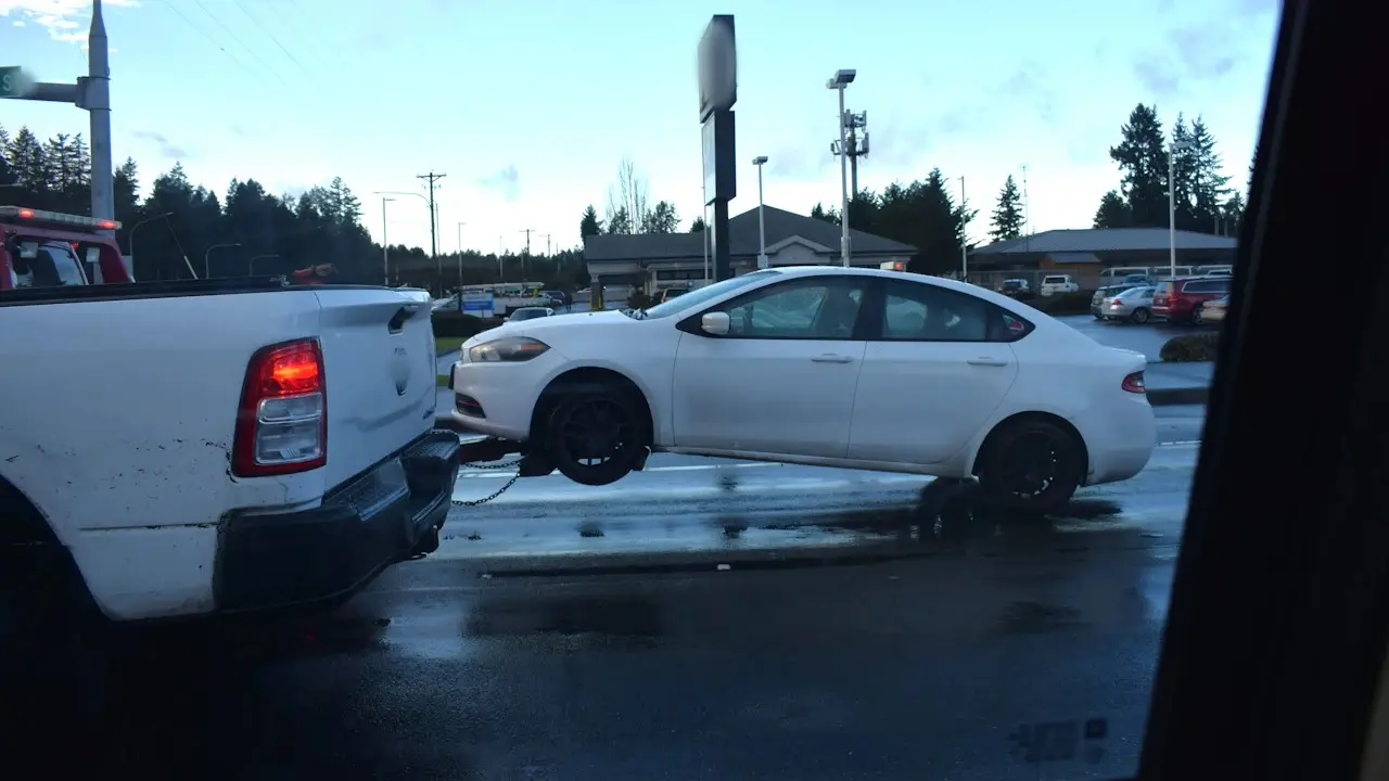 A white sedan towed by its front wheels behind a pickup, seen through a car window.