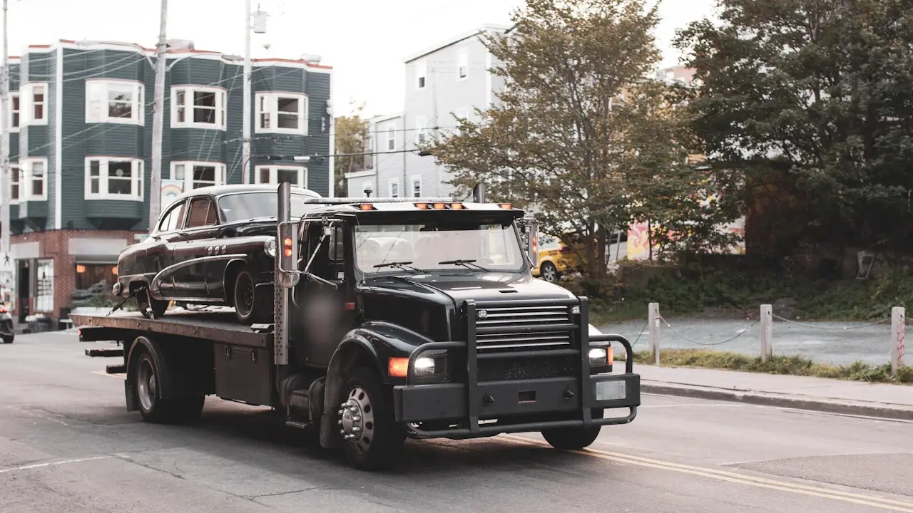 A tow truck carrying a classic black sedan on its flatbed down a city street.