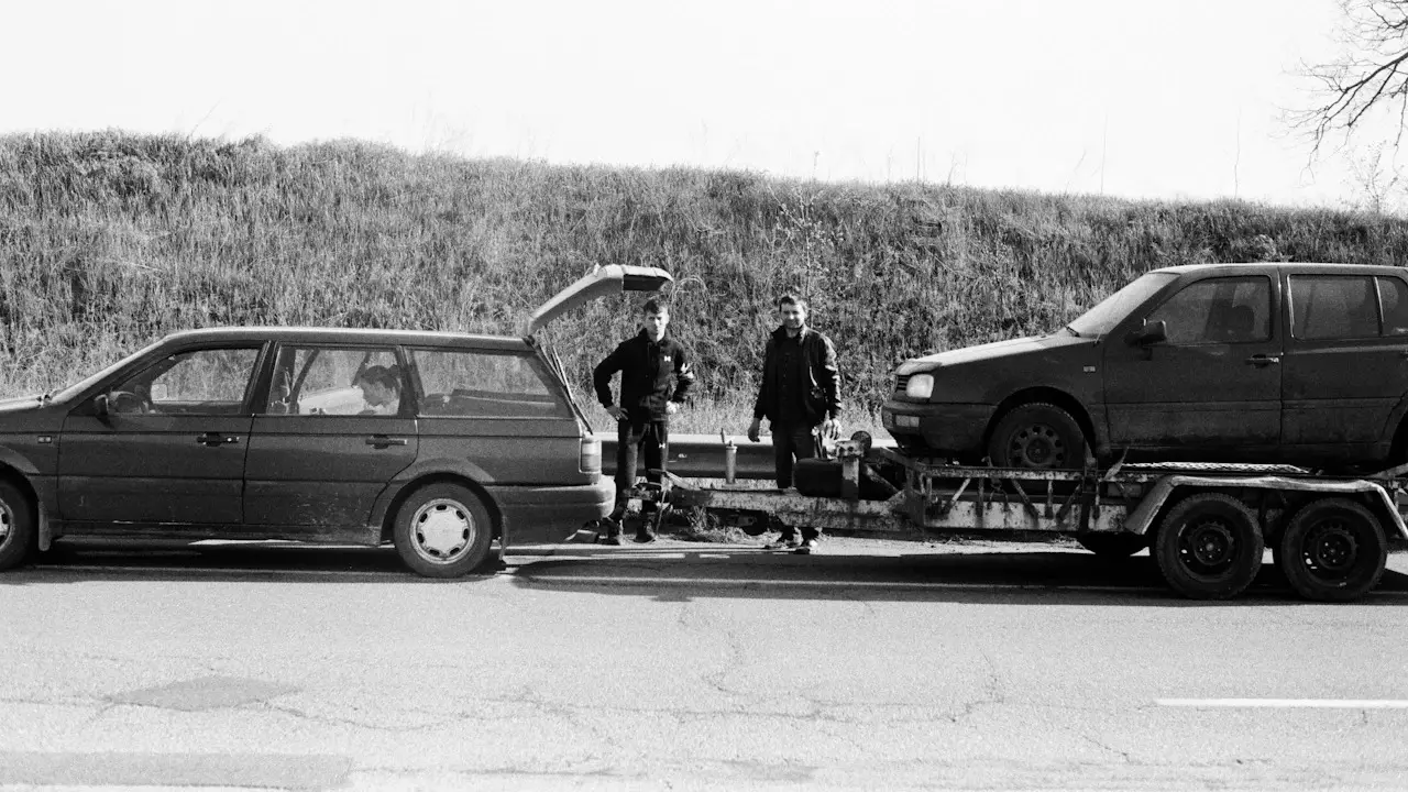 Black-and-white photo of two people beside a car on a flatbed trailer behind an estate car at a roadside.