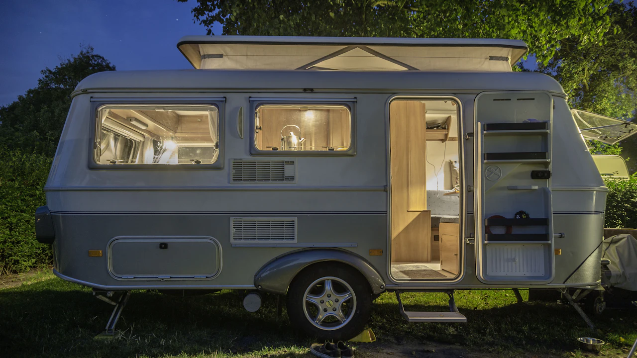 A small pale caravan on a grassy pitch after dark, its door open and its interior lit, a kettle visible through the side window