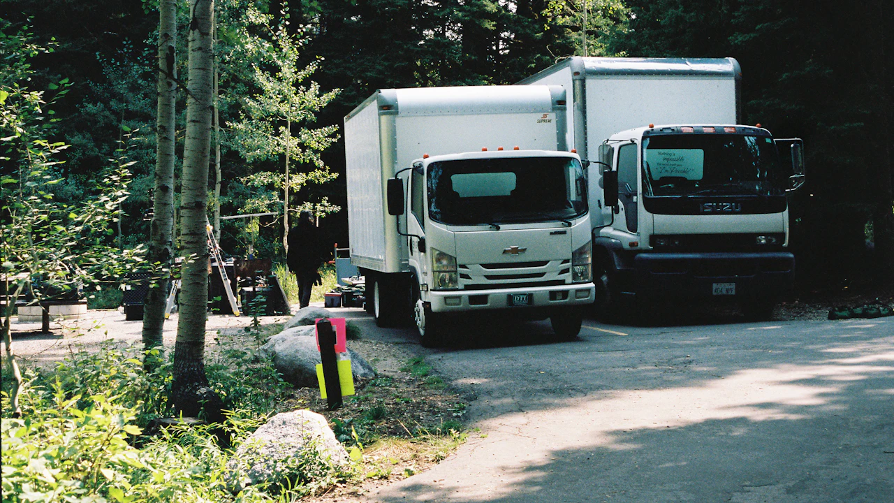 Two white cab-over box trucks parked nose-out on a gravel clearing among tall conifers. The nearer one carries a Chevrolet bowtie on its grille; the one beside it is badged Isuzu. Both have square cargo bodies considerably taller than their cabs.