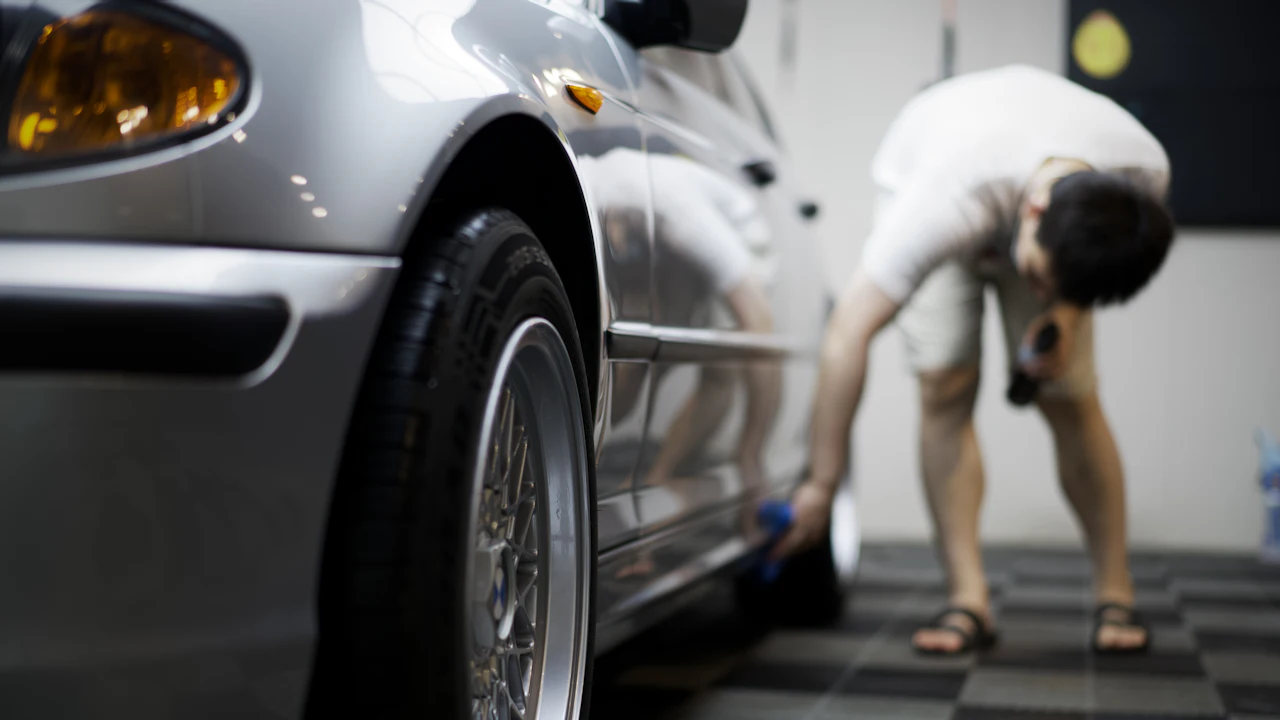 A car raised in a workshop with a wheel in the foreground and a technician working behind it