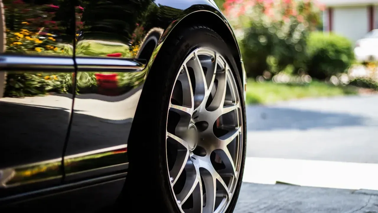 A black sedan's front wheel and low-profile tyre beside a kerb, with multi-spoke alloy rims and the car's glossy flank reflecting a garden.