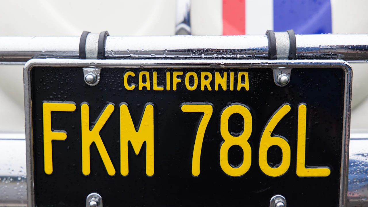 A black-and-yellow California licence plate photographed close up in the rain, bolted into a chrome frame on a pale vehicle