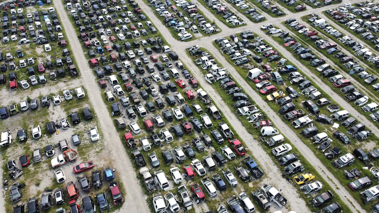 A drone view of a salvage yard, several hundred wrecked and stripped cars laid out in long rows across gravel and grass