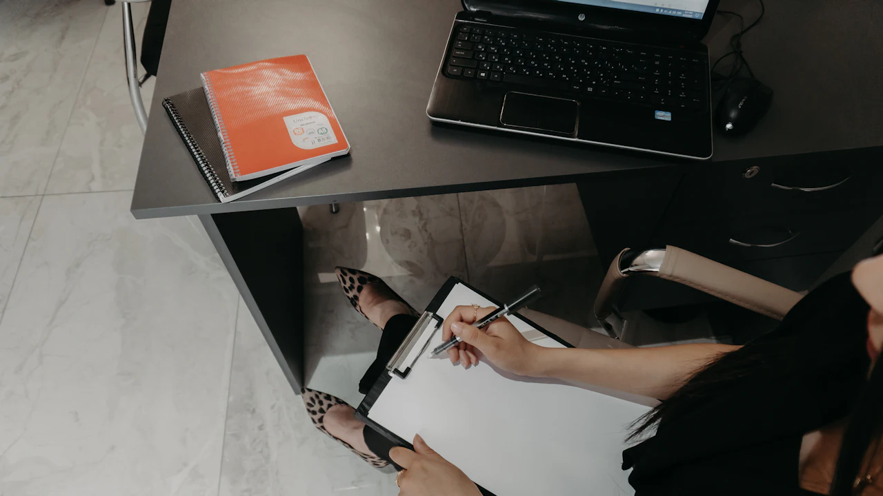 Overhead view of a person at a dark desk writing on a clipboard, an orange notebook and an open laptop beside them on the desktop.