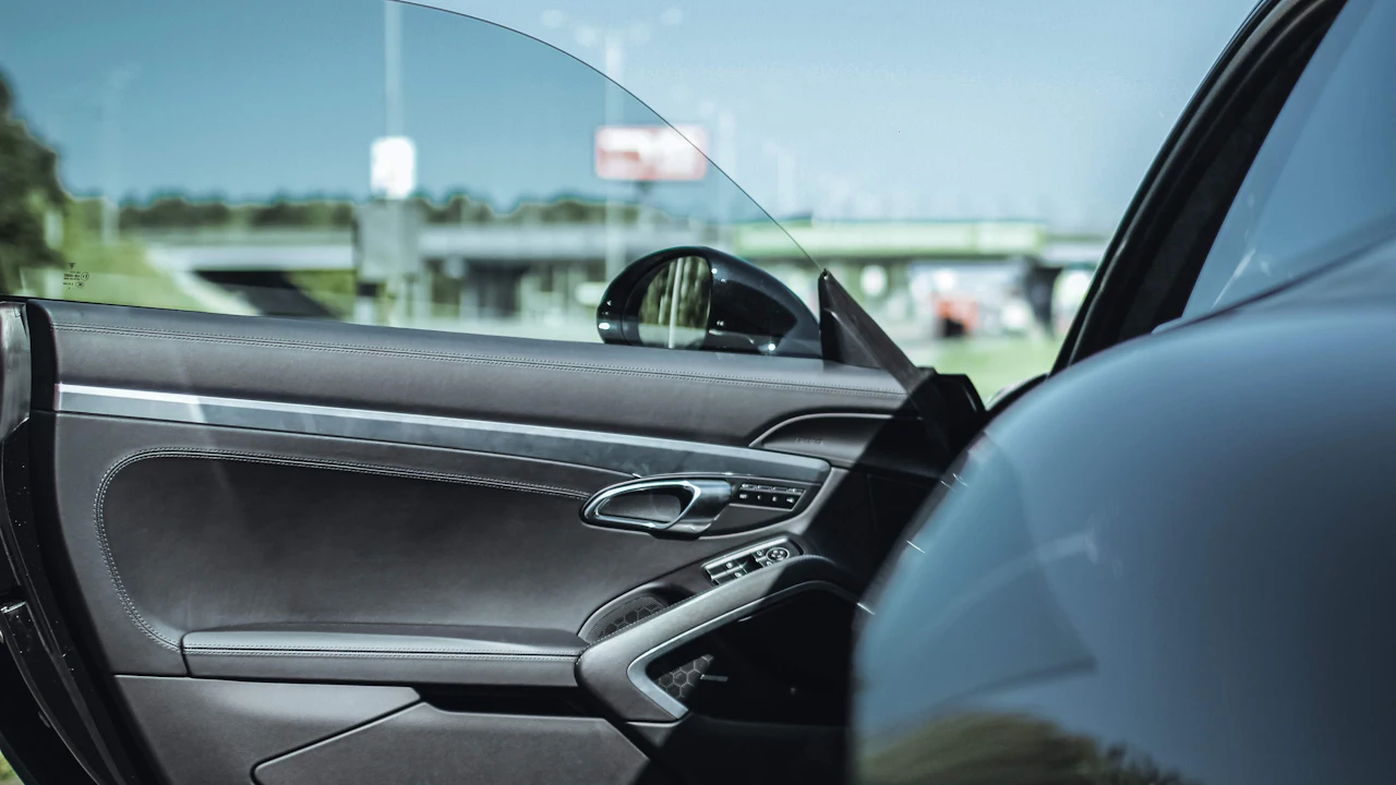 The open driver's door of a car, the door panel and interior on the left and the body pillar and window glass on the right, a car park visible beyond.