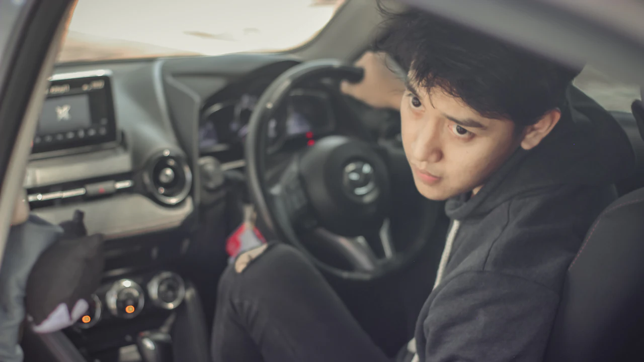 A young person in a car's driver seat looking over their shoulder, the dashboard and steering wheel beside them