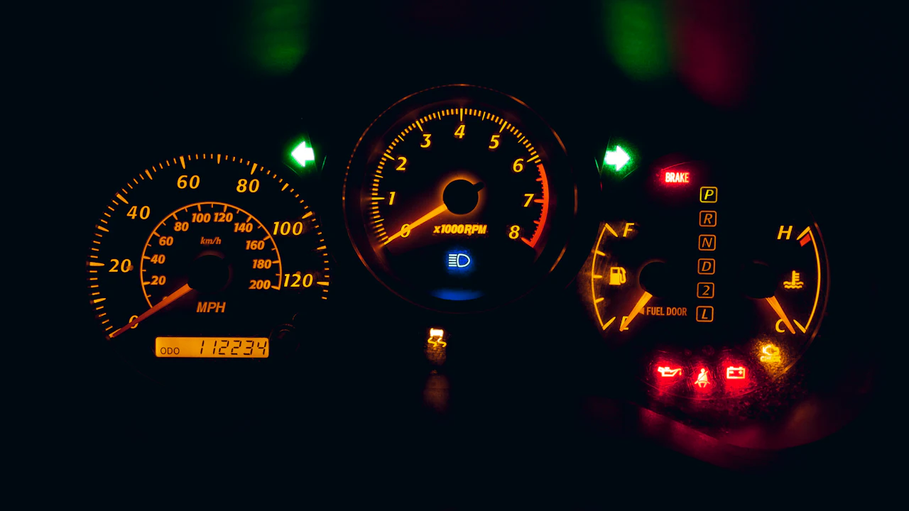 A car instrument cluster photographed in the dark, speedometer and rev counter glowing amber with a row of red and green warning symbols lit beside them