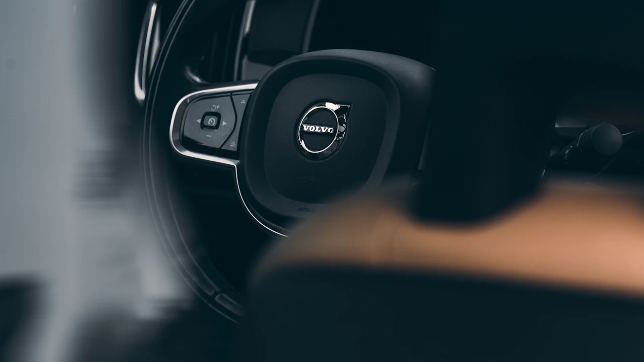 A Volvo steering wheel photographed close in low light, the badge sharp at the centre and a tan leather seat behind