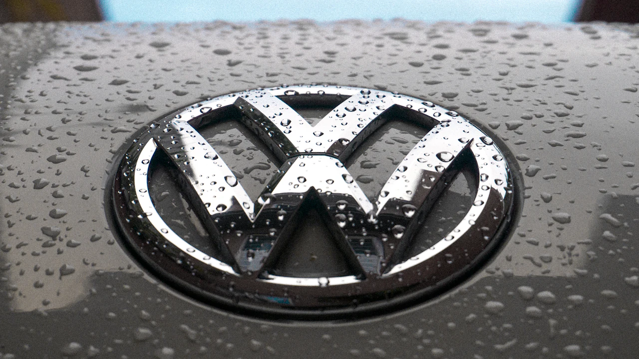 A chrome Volkswagen roundel on a grey bonnet, photographed close and filling most of the frame, the paint around it beaded with rain.
