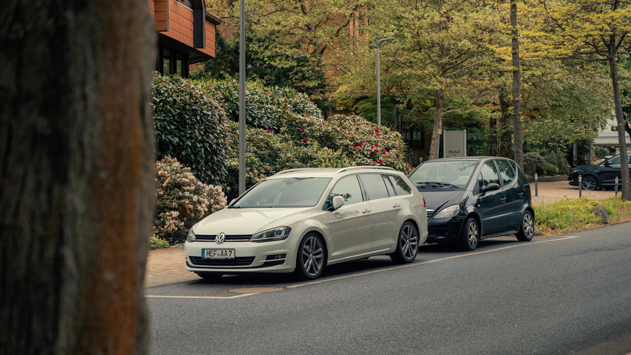 A white estate car parked at the kerb of a leafy residential street, a small dark hatchback behind it and autumn trees above
