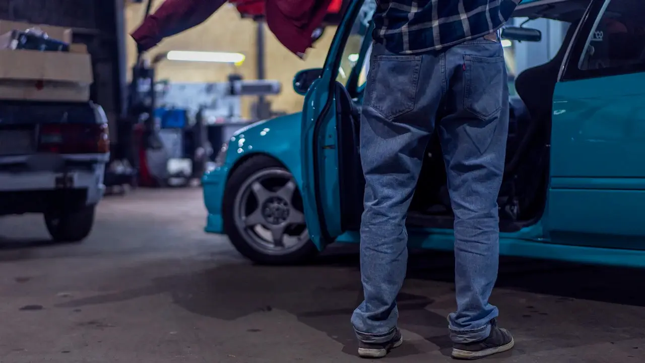 A person standing by a teal car's open door in a workshop.