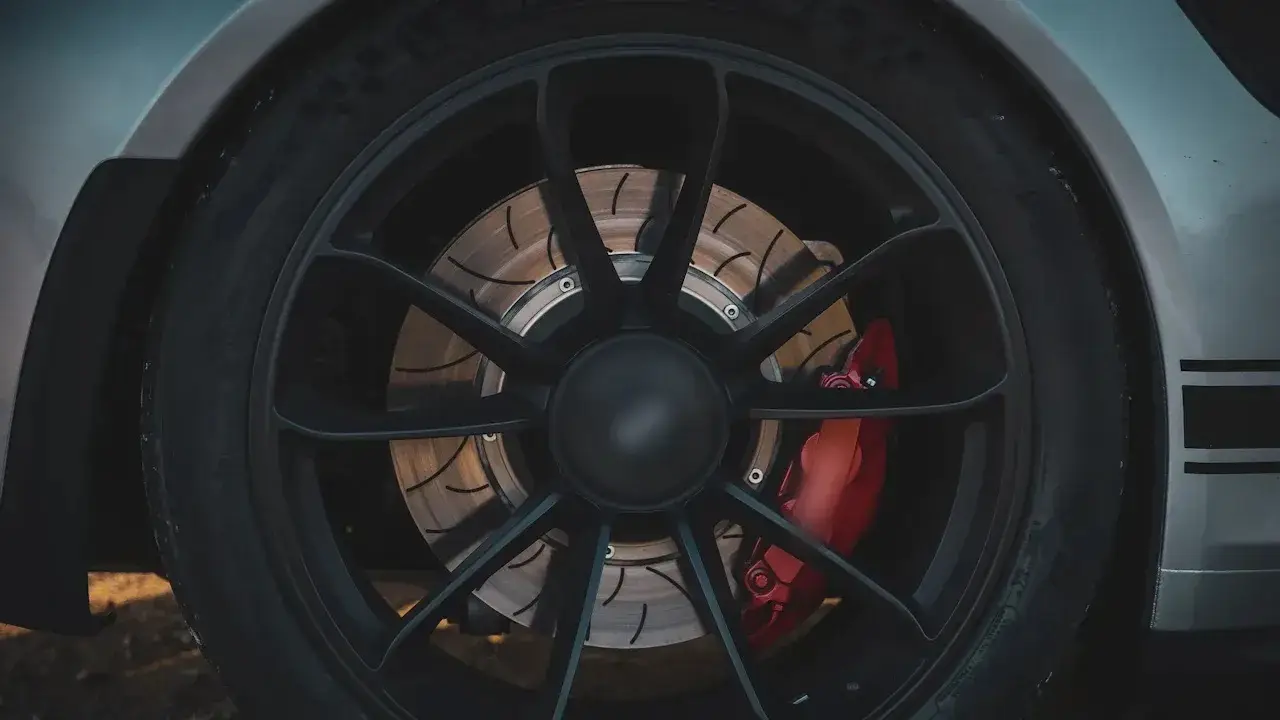 A black ten-spoke wheel on a silver car, with the slotted brake disc and a red brake caliper visible through the spokes.