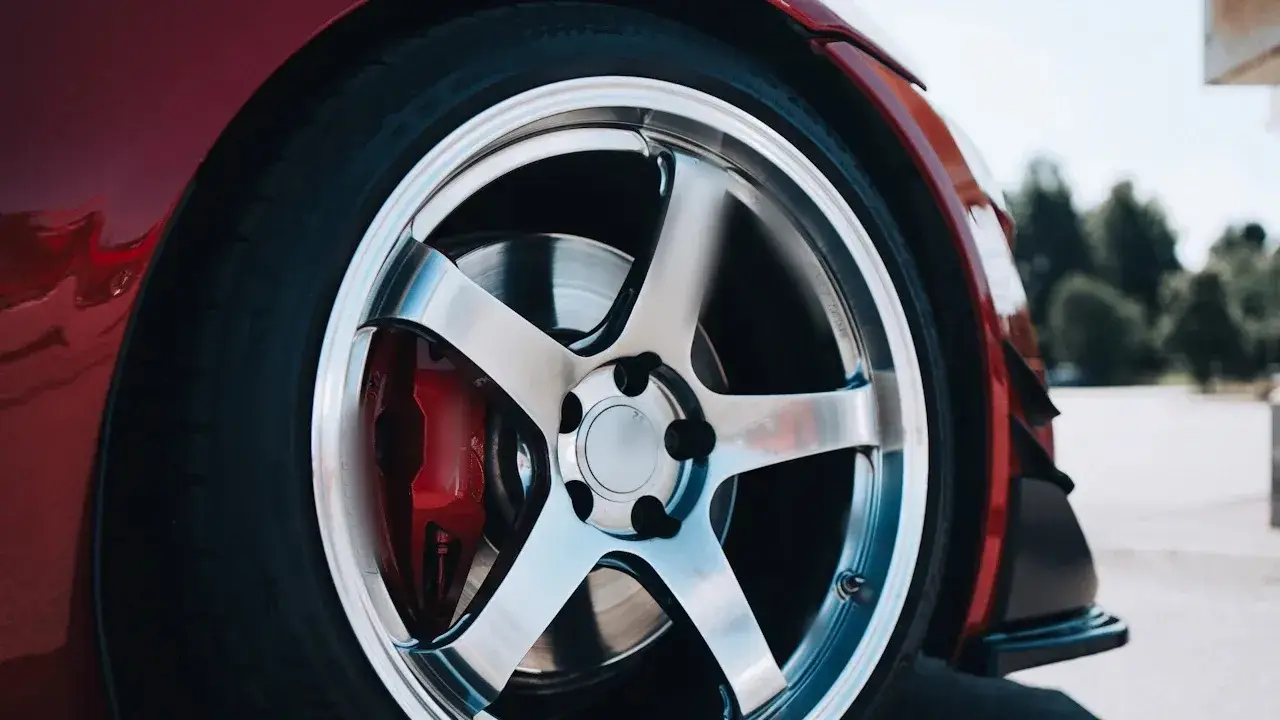 A close-up of a car's silver five-spoke wheel on a red car, with the red brake caliper, the brake rotor and the lug nuts visible through the spokes.